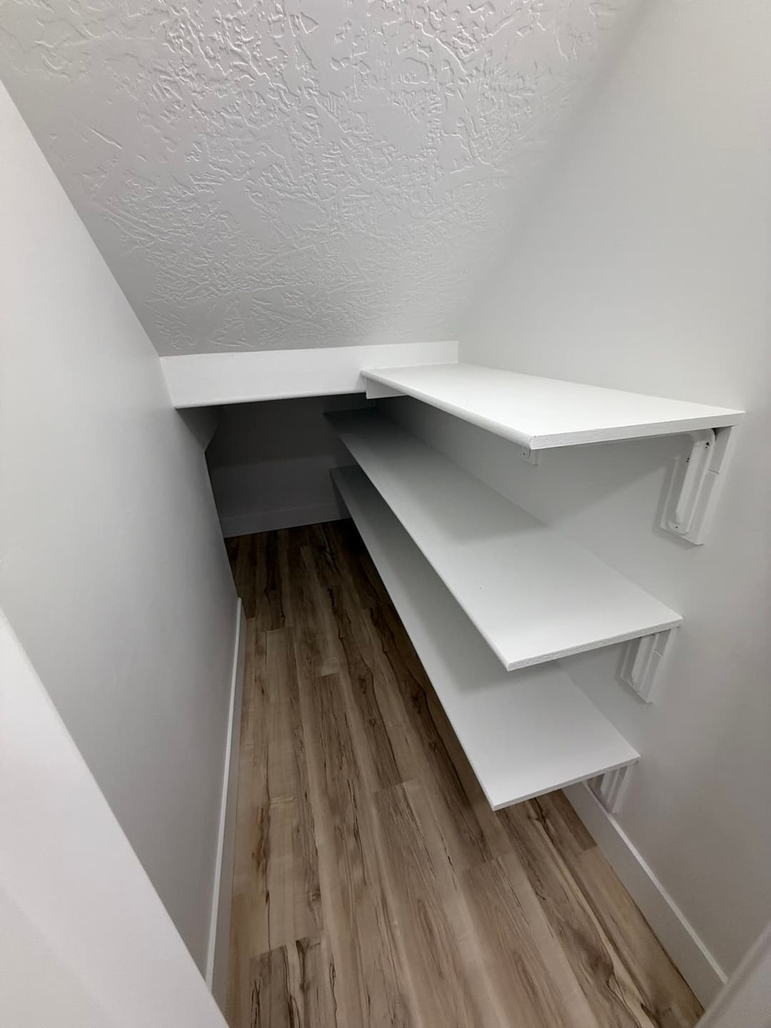 White shelving units in a small, angled closet with a light wood floor.