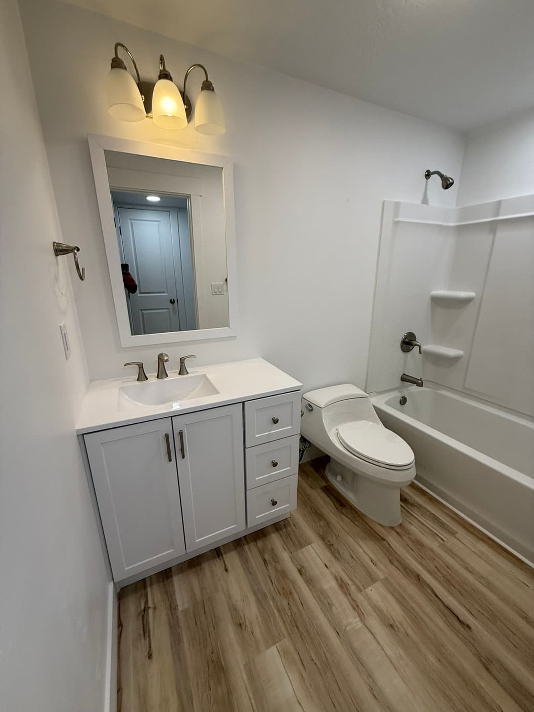 Modern bathroom with a white vanity, mirror, shower-tub combo, and wood-style flooring.