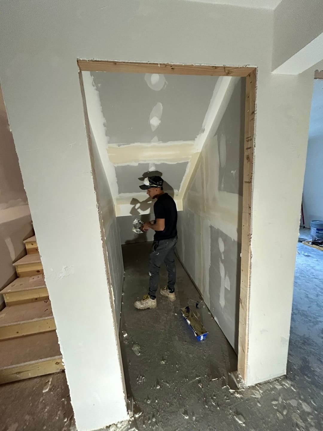 Person applying drywall compound in a hallway under renovation with unfinished stairs visible.
