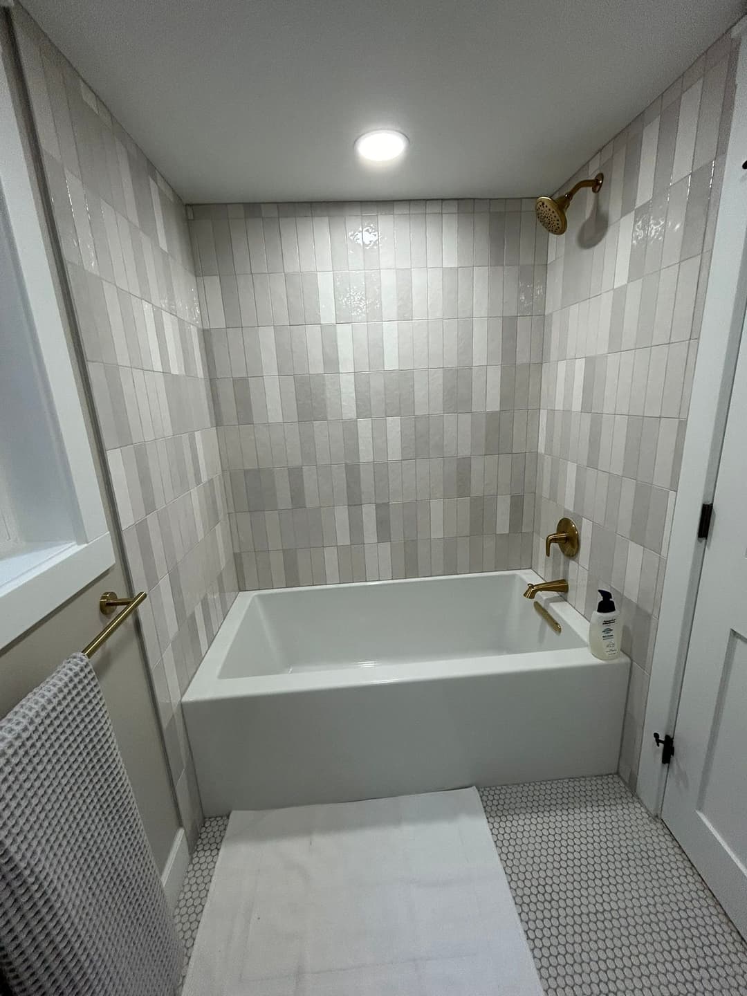 Modern bathroom with gray tile walls, a white bathtub, gold fixtures, and a plush rug.