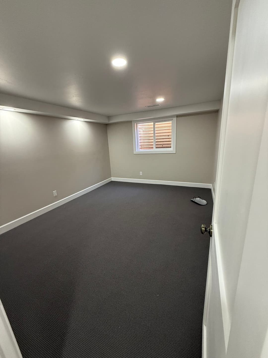 Empty basement room with gray carpet and natural light from a window, ready for renovation.